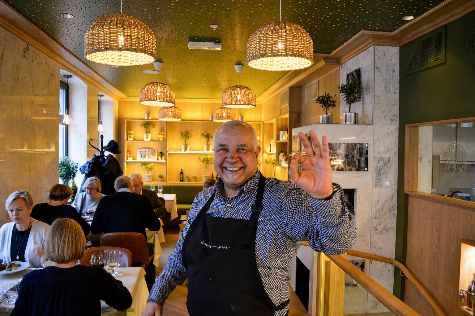 Verten i sort forkle med brodert logo står midt i salen og vinker mot kamera